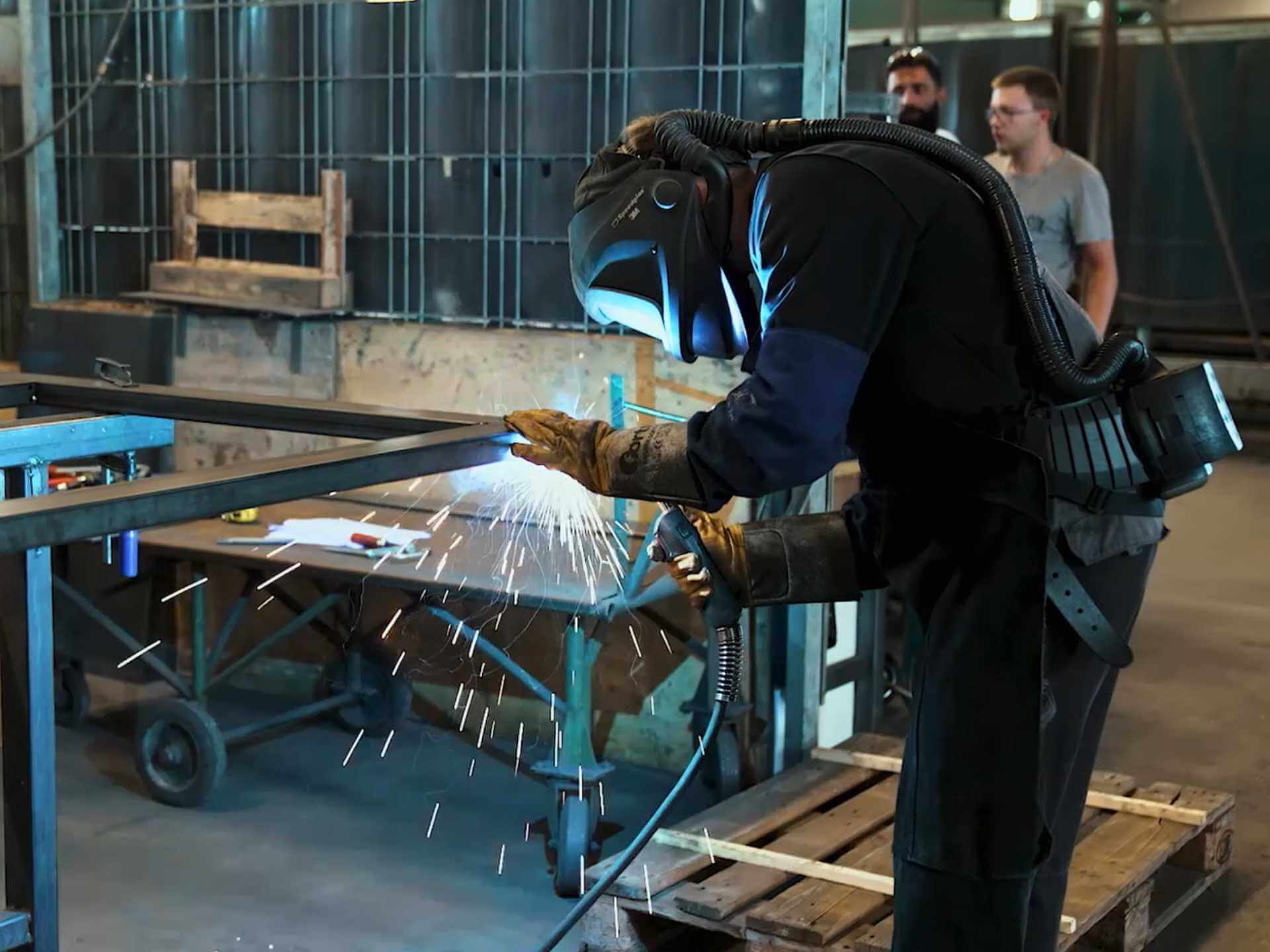Ein Mann mit Schweißmaske und Schutzkleidung schweißt ein Metallgerüst. Die Funken sprühen weißblau in der dunklen Werkshalle.