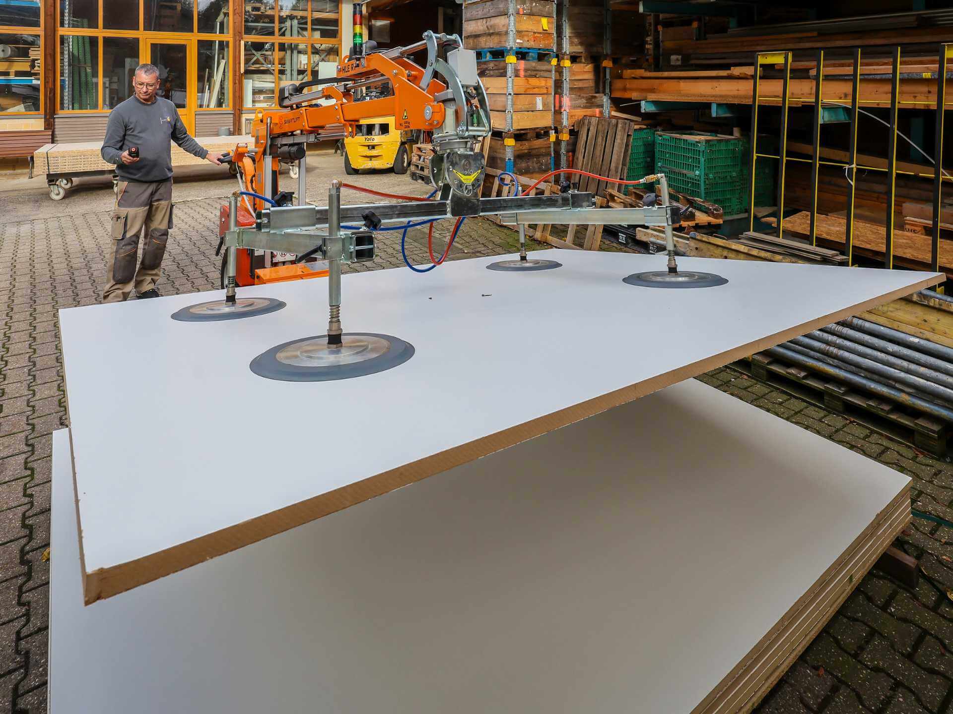 Ein Mann Ende 50 bedient einen Robotor. Das Gerät hebt mittels vier großer Saugscheiben eine schwere Glasplatte an.