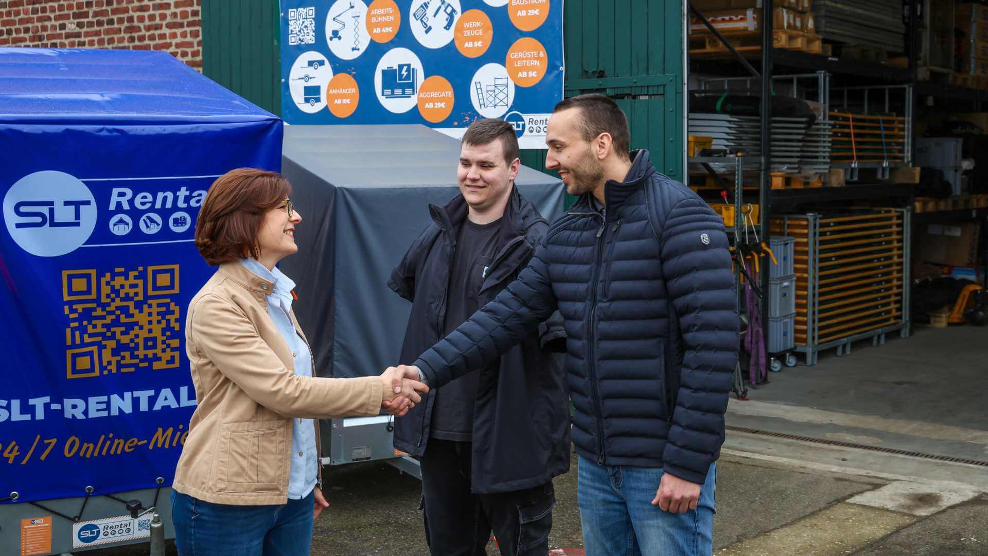 Eine Frau begrüßt einen Mann mittleren Alters auf einem Gewerbehof mit Handschlag. Zwischen ihnen steht ein junger Mann, dem beide Arme fehlen.