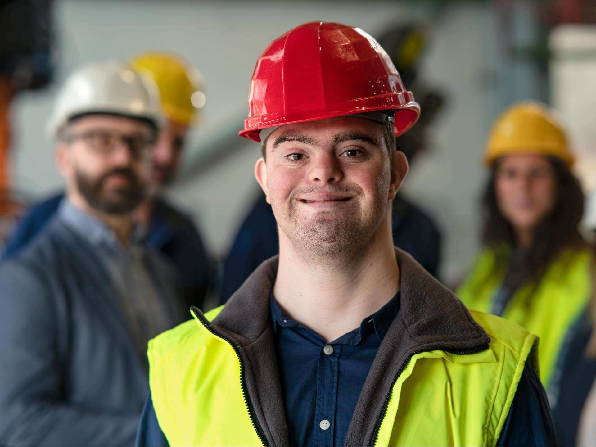 Ein junger Mann mit Downsyndrom trägt einen roten Schutzhelm und eine gelbe Warnweste. Er lächelt. Im Hintergrund ist verschwommen eine Gruppe von Männern und Frauen vor einem Lagerregal zu sehen. Auch sie tragen Helme und Schutzwesten.