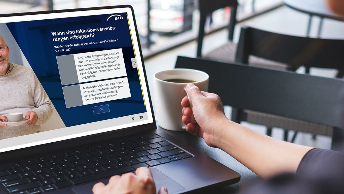 Auf einem Laptop-Monitor ist eine Szene aus dem Selbstlernkurs „Inklusionsvereinbarung“ zu sehen. Ein älterer Herr stellt Fragen, die angeklickt werden können.